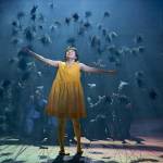 Satsuki (Ami Okumura Jones) wearing a yellow dress and holding her arms out as the Soot Sprites go around and behind her.